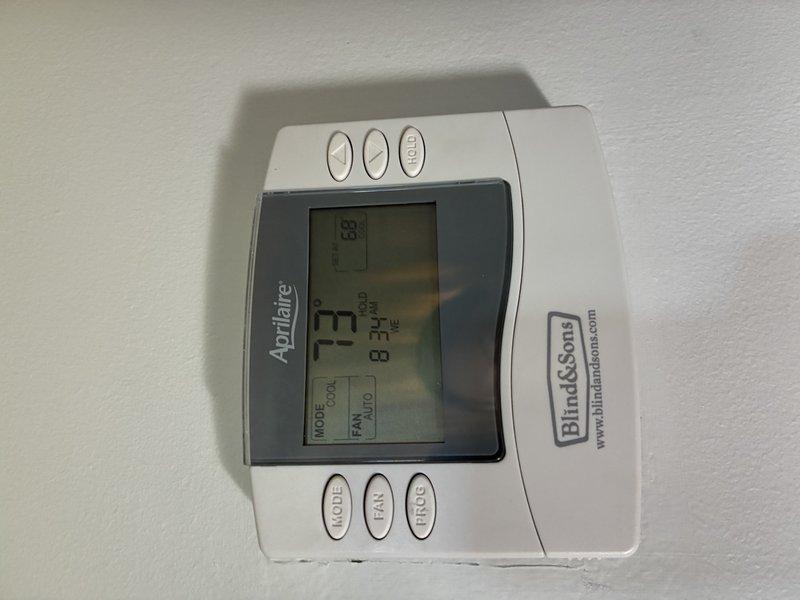 Responded to service call for non-functioning Aprilaire thermostat following recent PCB installation. Inspected thermostat wiring connections at wall-mounted control device, verified proper wire terminations at numbered terminal block, and confirmed thermostat display was operational showing 73° with system in cooling mode and fan set to auto. Documented wiring configuration and thermostat functionality with photos for service records.