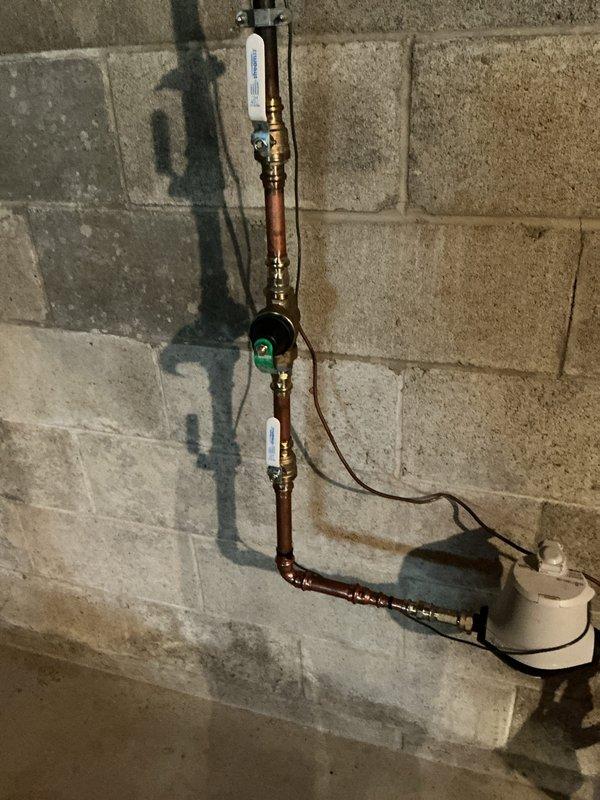 Installed new automatic shutoff valve assembly on main water line in basement of 1960s-era home where no existing valve was previously present. The installation included copper piping with brass fittings mounted to concrete block wall, incorporating a pressure monitoring gauge and white cylindrical reservoir component connected via tubing. Exterior hose bib confirmed operational upon completion of interior valve installation.