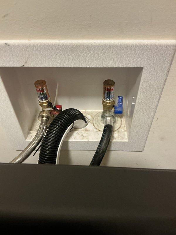 Responded to service call for laundry room plumbing issues involving lack of hot water supply and water infiltration into HVAC vents during washer operation. Upon inspection, identified compromised water supply connections at the wall-mounted outlet box serving the washing machine, including failed hot water valve assembly and deteriorated supply line connections causing water discharge into adjacent ventilation system. Documented existing conditions showing wall-mounted plumbing box with brass shut-off valves, braided stainless steel flex line, and rubber supply hoses with visible water staining at connection points, as well as ceiling vent location where water intrusion occurred.