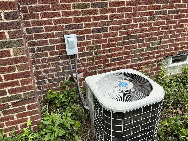 Conducted on-site evaluation of existing outdoor air conditioning system for replacement estimate. Documented current cylindrical condenser unit positioned against brick exterior wall, noting electrical disconnect box, refrigerant line routing, and equipment accessibility. Assessed surrounding landscape conditions and clearance requirements for future equipment installation.