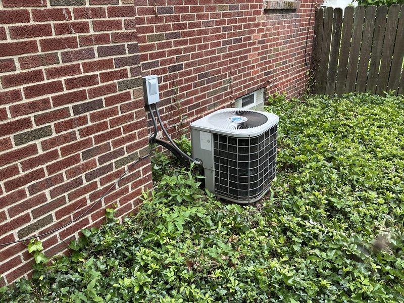 Conducted on-site evaluation of existing outdoor air conditioning system for replacement estimate. Documented current cylindrical condenser unit positioned against brick exterior wall, noting electrical disconnect box, refrigerant line routing, and equipment accessibility. Assessed surrounding landscape conditions and clearance requirements for future equipment installation.