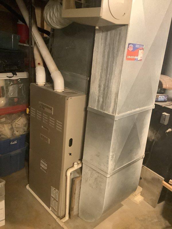 Conducted on-site evaluation of 25-year-old residential furnace system located in basement utility area for equipment replacement estimate. Assessed existing beige furnace unit with white PVC venting, adjacent sheet metal ductwork assembly, and associated mechanical components. Documented current system configuration including vertical ductwork sections, PVC piping connections, and overall equipment condition through photographic documentation to support replacement proposal development.