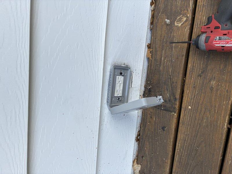 Corrected exterior electrical outlet installation that was previously mounted in improper orientation. Verified weatherproof GFCI outlet box and conduit are now installed to code specifications on building exterior where white siding meets wooden planks. Tested outlet functionality and confirmed proper grounding and weather protection.