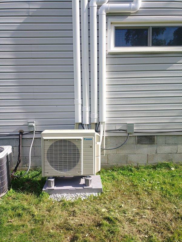Completed installation of a 5-head mini-split system with Daikin outdoor condenser unit mounted on concrete pad with stone base. Installed refrigerant line sets with white PVC line covers running vertically along exterior siding, electrical disconnect box, and all associated condensate drainage piping to manufacturer specifications.