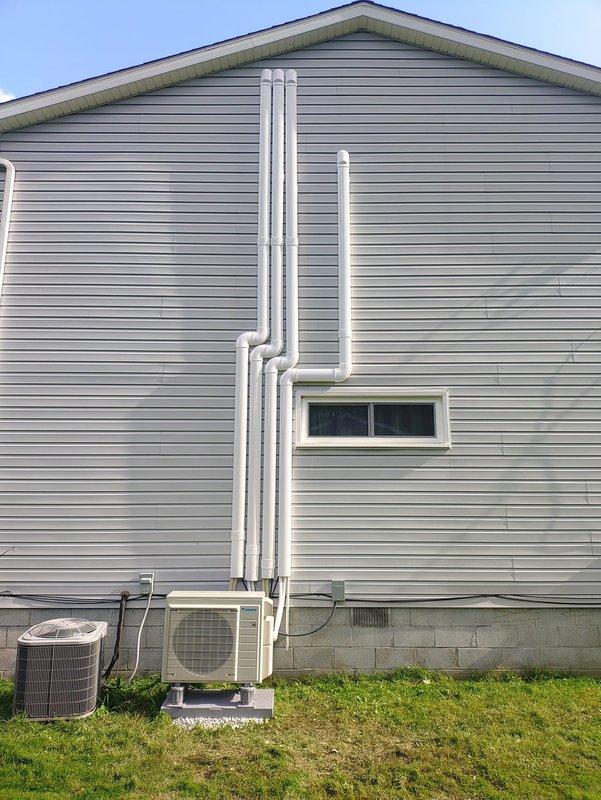 Completed installation of a 5-head mini-split system with Daikin outdoor condenser unit mounted on concrete pad with stone base. Installed refrigerant line sets with white PVC line covers running vertically along exterior siding, electrical disconnect box, and all associated condensate drainage piping to manufacturer specifications.