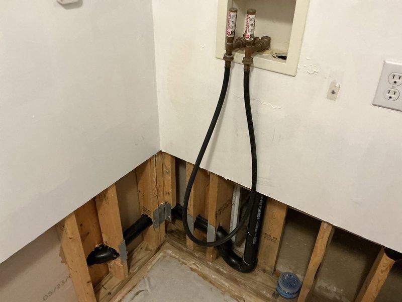 Responded to a plumbing service call for a water leak in the laundry room and a clogged toilet that were initially addressed in May. Upon arrival, inspected the area where drywall had been removed to expose wall studs and plumbing infrastructure. Identified water supply line connections requiring attention, with copper fittings and pressure gauges visible in the wall box. Assessed black flexible supply hoses running from floor level to upper wall connections, along with copper pipe fittings within the exposed cavity. Noted water staining on concrete floor indicating previous leak activity. Cleared the clogged toilet to restore proper drainage function. Documented the exposed plumbing configuration showing supply line connections with red and white identification tags, along with visible drainage components in the lower wall section. The leak repair was completed to allow restoration work to proceed, with all supply connections properly secured and tested for integrity.