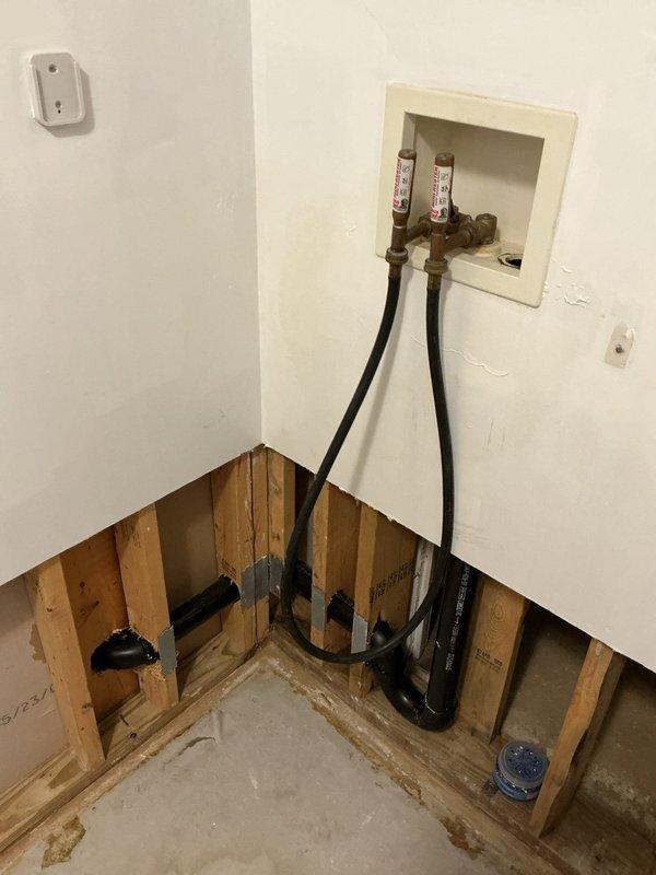 Responded to a plumbing service call for a water leak in the laundry room and a clogged toilet that were initially addressed in May. Upon arrival, inspected the area where drywall had been removed to expose wall studs and plumbing infrastructure. Identified water supply line connections requiring attention, with copper fittings and pressure gauges visible in the wall box. Assessed black flexible supply hoses running from floor level to upper wall connections, along with copper pipe fittings within the exposed cavity. Noted water staining on concrete floor indicating previous leak activity. Cleared the clogged toilet to restore proper drainage function. Documented the exposed plumbing configuration showing supply line connections with red and white identification tags, along with visible drainage components in the lower wall section. The leak repair was completed to allow restoration work to proceed, with all supply connections properly secured and tested for integrity.