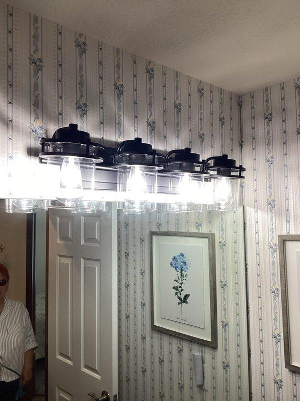Installed two new vanity light fixtures in separate bathrooms. The first bathroom received a modern four-light fixture with black metal construction and clear glass shades mounted above the vanity. The second bathroom was fitted with a traditional four-light fixture featuring brass-toned curved arms with white frosted glass shades, also positioned above the mirror and vanity area. Both fixtures were wired, mounted securely to the wall, tested for proper operation, and confirmed to be functioning correctly upon completion.