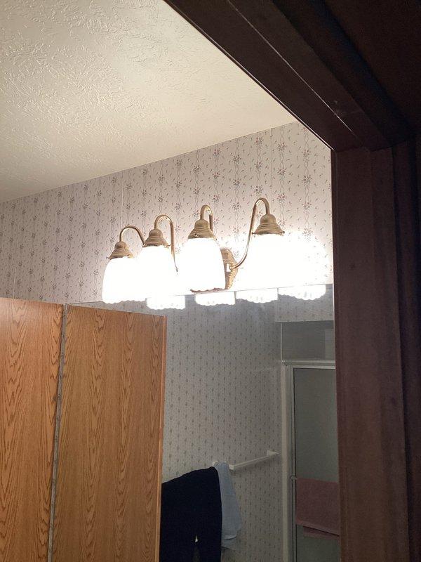 Installed two new vanity light fixtures in separate bathrooms. The first bathroom received a modern four-light fixture with black metal construction and clear glass shades mounted above the vanity. The second bathroom was fitted with a traditional four-light fixture featuring brass-toned curved arms with white frosted glass shades, also positioned above the mirror and vanity area. Both fixtures were wired, mounted securely to the wall, tested for proper operation, and confirmed to be functioning correctly upon completion.