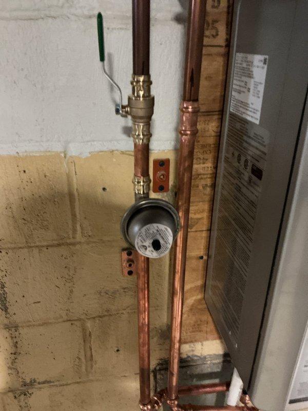 Completed tankless water heater installation in utility room. Installed new copper supply piping with brass ball valve and isolation valves, integrated flow meter with wall-mounted backing board, and connected unit to existing plumbing system. Modified drainage system with new PVC P-trap configuration and rubber coupling adapters to accommodate condensate discharge requirements. All connections tested and verified operational.