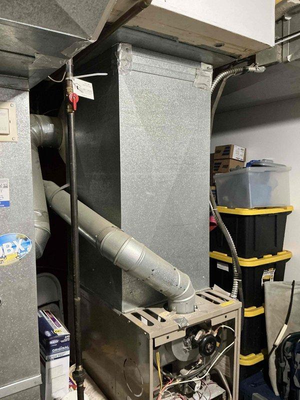 Conducted comprehensive evaluation of 29-year-old HVAC system and provided replacement estimate exceeding $7,000 for complete system upgrade. Documented existing basement air handler installation with metallic finish, black metal plenum, and associated ductwork connections, noting system age and operational status. Photographed mechanical room layout including water heater and electrical components to support replacement proposal specifications.