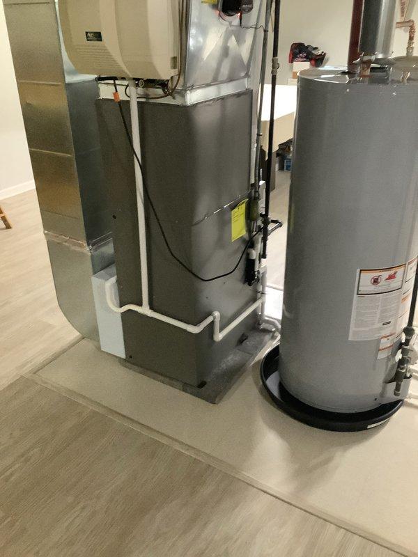 Completed installation of free-standing dehumidifier unit in residential mechanical room to improve indoor air quality. Unit was positioned on protective mat alongside existing HVAC furnace and water heater, with white PVC condensate drainage line properly routed and connected to ensure efficient moisture removal and system operation.