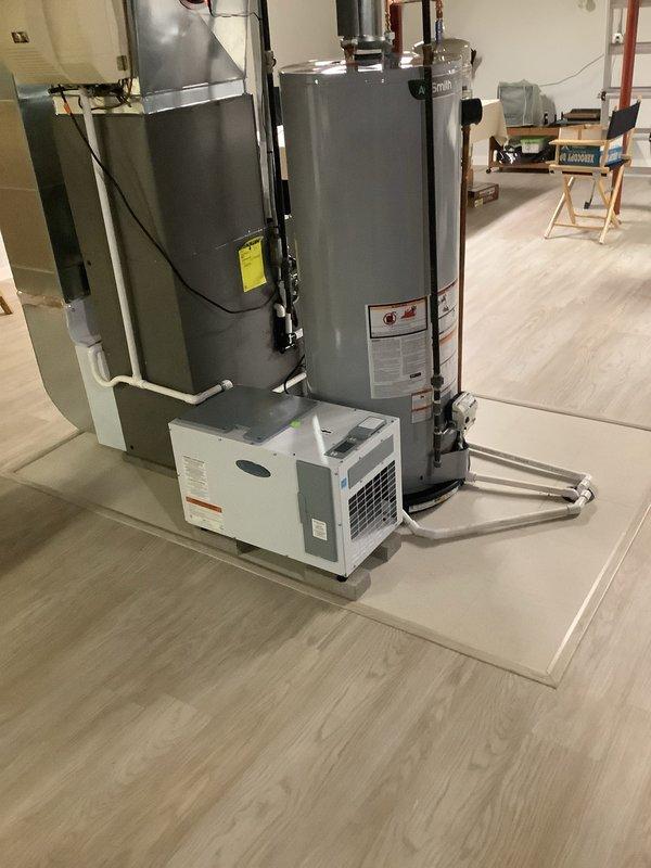 Completed installation of free-standing dehumidifier unit in residential mechanical room to improve indoor air quality. Unit was positioned on protective mat alongside existing HVAC furnace and water heater, with white PVC condensate drainage line properly routed and connected to ensure efficient moisture removal and system operation.