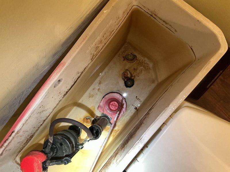 Responded to reported "toilet rain" issue at 20-year-old residential property. Upon arrival, removed toilet tank lid and conducted internal inspection of flush mechanism components. Found aged internal parts with significant mineral deposits, rust staining, and discoloration throughout tank interior. Identified deteriorated flapper valve, fill valve assembly with float ball, and flush handle mechanism showing signs of wear consistent with original installation age. Documented existing conditions of tank components including corrosion and mineral buildup affecting proper operation. Photographed toilet bowl with cleaning product present and complete tank interior showing all mechanical components requiring attention for proper flush cycle operation.