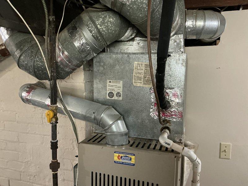 Conducted on-site evaluation of non-functional HVAC system with outdoor air conditioning unit and indoor furnace, both exceeding 20 years of service life. System was blowing warm air only, indicating compressor failure or refrigerant loss in the outdoor condensing unit. Documented existing equipment configuration including beige outdoor AC unit with cylindrical fan grille positioned on ground-level pad with electrical conduit and refrigerant lines running to building, and indoor furnace installation with air handler in basement utility area showing significant dust accumulation on components and ductwork. Provided comprehensive replacement estimate for complete HVAC system upgrade to address failed cooling capacity and aging equipment.