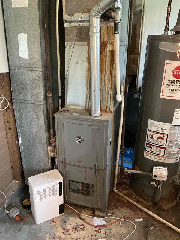 Completed comprehensive assessment of existing HVAC system for replacement estimate at residential property. Evaluated aging outdoor air conditioning condenser unit with cylindrical design and black wire mesh grille, showing over 10 years of service life with signs of weathering and vegetation encroachment around base. Inspected indoor furnace unit located in basement mechanical room, noting rust staining on exterior surfaces and evidence of previous moisture issues in surrounding area. Documented current system configuration including water heater, dehumidifier, and associated ductwork and piping. Photographed all equipment and conditions to support replacement recommendations for new AC system installation.