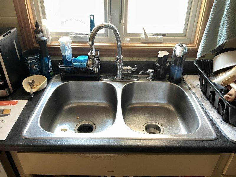 Replaced existing kitchen sink and faucet assembly per customer request. Removed double-basin stainless steel sink with chrome dual-handle faucet and side sprayer, then installed new single-bowl black sink with corresponding fixtures. Completed all necessary plumbing connections under cabinet, ensured proper drainage function, and verified no leaks in water supply lines or drain assembly.