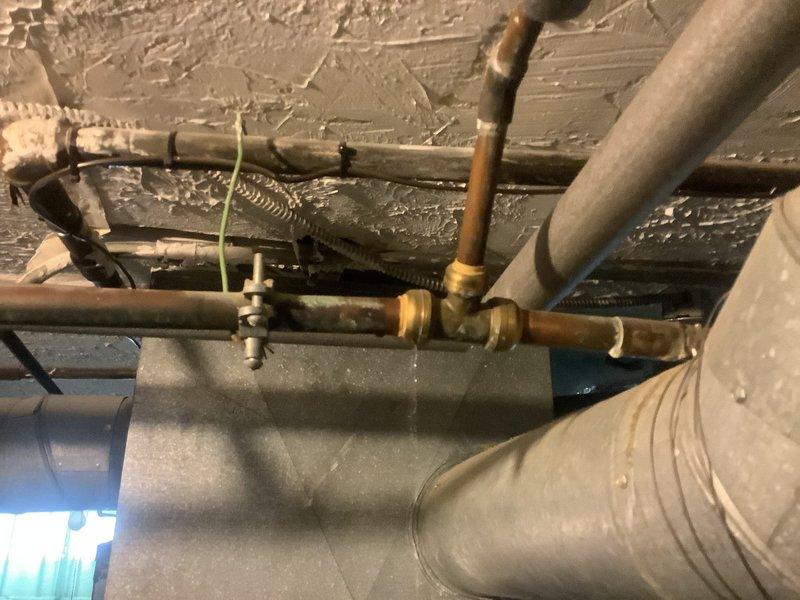 Responded to water leak complaint near recently installed flue pipe. Upon inspection of basement mechanical room, identified active leak at copper pipe connection point adjacent to ductwork. Leak originated from compromised joint near T-fitting configuration where copper piping interfaces with main flue assembly. Assessed surrounding piping infrastructure including insulated lines and galvanized steel components to determine extent of water damage and verify no additional leak sources present. Documented existing conditions showing aged plumbing system with oxidation on copper fittings and deteriorated ceiling surfaces indicating previous moisture exposure. Tightened affected connection and verified proper seal at joint. Monitored system under pressure to confirm leak resolution and no further water discharge from repair area.
