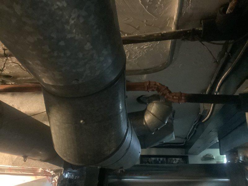 Responded to water leak complaint near recently installed flue pipe. Upon inspection of basement mechanical room, identified active leak at copper pipe connection point adjacent to ductwork. Leak originated from compromised joint near T-fitting configuration where copper piping interfaces with main flue assembly. Assessed surrounding piping infrastructure including insulated lines and galvanized steel components to determine extent of water damage and verify no additional leak sources present. Documented existing conditions showing aged plumbing system with oxidation on copper fittings and deteriorated ceiling surfaces indicating previous moisture exposure. Tightened affected connection and verified proper seal at joint. Monitored system under pressure to confirm leak resolution and no further water discharge from repair area.