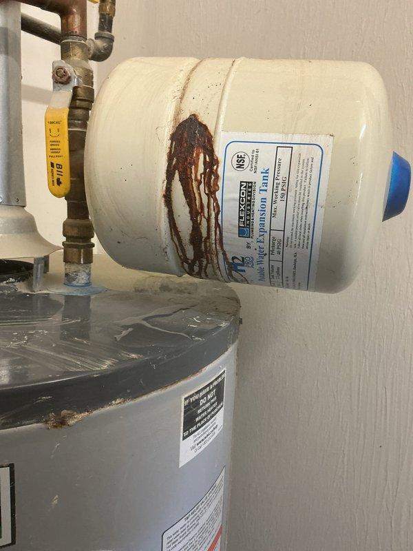 # Water Heater Expansion Tank Leak Repair

Responded to a water leak complaint involving the expansion tank mounted above the water heater. Upon inspection, confirmed active leaking from the thermal expansion tank connection point. The expansion tank exhibited significant rust-colored staining and corrosion streaking down the front surface, indicating prolonged moisture exposure from the failed connection or tank integrity compromise.

Documented the condition of the complete water heating system, including the expansion tank, copper supply piping, shut-off valves, and main water heater unit. The rust deterioration pattern originated from the upper pipe connection area where the expansion tank interfaces with the copper plumbing assembly. The corrosion and staining suggested the leak had been present for an extended period, allowing mineral deposits and oxidation to accumulate on the tank surface.

Photographed the installation showing the horizontal-mounted cream-colored expansion tan