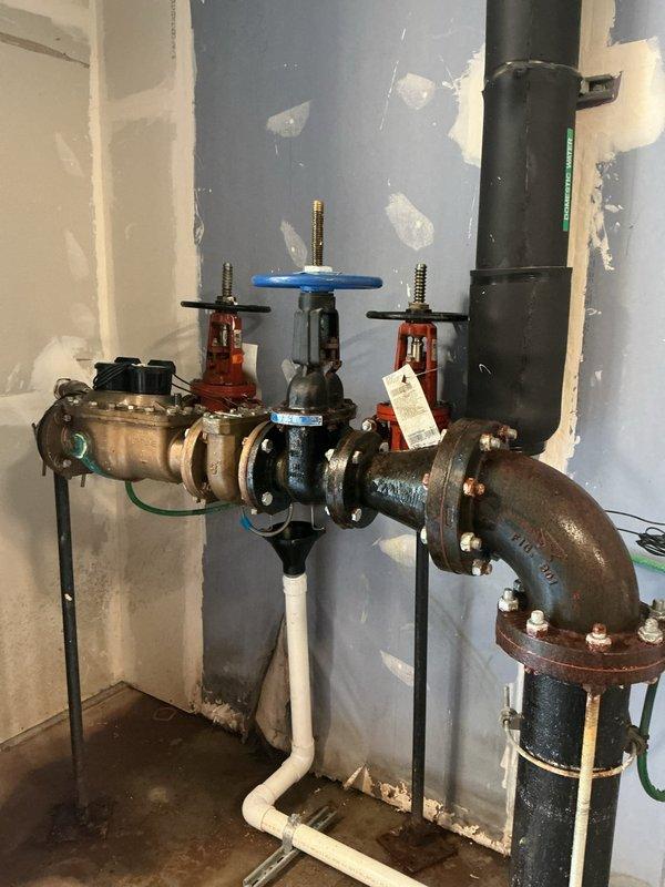 Responded to main line leak in unit 207 where customer had already opened wall access. Accessed plumbing assembly through existing cutout revealing corroded piping with active leak at valve connections. Replaced deteriorated main line section including installation of new brass fittings, shutoff valves with black, blue, and red handles for system isolation, and flanged connections. Addressed significant rust and corrosion on existing elbow sections. Integrated new PEX supply lines (blue tubing) with existing copper and galvanized steel piping. Installed pressure regulation components and ensured proper valve operation for future maintenance access. Protected toilet fixture during work and verified system integrity with pressure testing before closing.