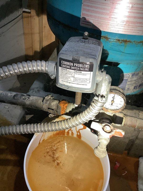 Responded to service call for water softener leaking. Upon arrival, inspected plumbing configuration and identified active leak at corroded pipe fittings and connections beneath the well pressure tank system. Found significant rust staining on metal fittings and sediment-laden water collecting in bucket below the leak point. Assessed the water treatment system including the blue Well-Rite pressure tank, associated PVC piping, valves, and connections showing signs of age and oxidation. Documented equipment condition with photos showing the extent of corrosion on threaded connections and the compromised plumbing assembly requiring repair to stop the active water leak.