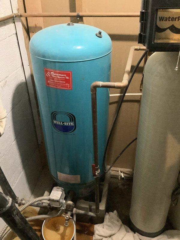 Responded to service call for water softener leaking. Upon arrival, inspected plumbing configuration and identified active leak at corroded pipe fittings and connections beneath the well pressure tank system. Found significant rust staining on metal fittings and sediment-laden water collecting in bucket below the leak point. Assessed the water treatment system including the blue Well-Rite pressure tank, associated PVC piping, valves, and connections showing signs of age and oxidation. Documented equipment condition with photos showing the extent of corrosion on threaded connections and the compromised plumbing assembly requiring repair to stop the active water leak.