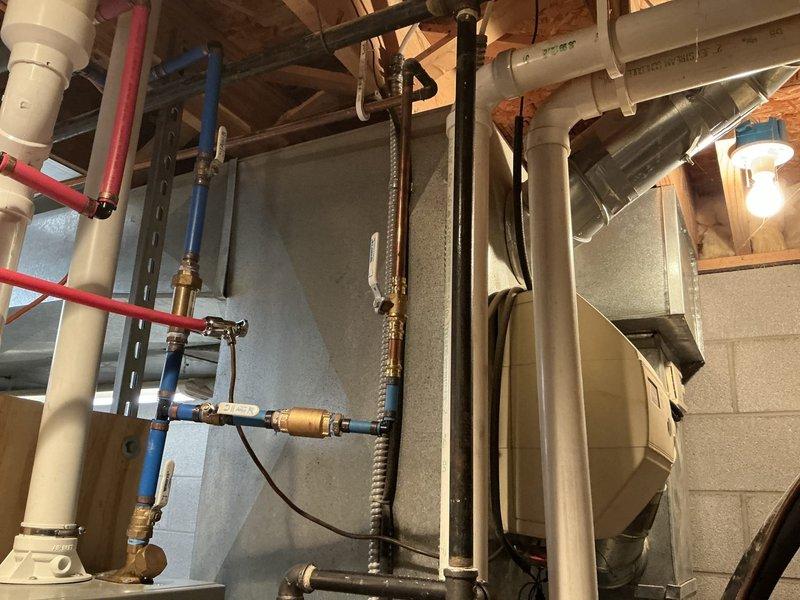 Provided estimate for replacement of 27-year-old furnace system in basement mechanical room. Existing gray rectangular furnace unit shows age-related wear with metallic ductwork extending from top and side, white PVC condensate lines, and flexible gas line connector with brass valve assembly. Documented current installation including plumbing lines (white PVC, red and blue PEX water lines with brass fittings), black metal strapping, and typical residential HVAC infrastructure in unfinished basement space with exposed wooden joists and concrete block walls.