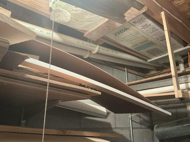Provided estimate for replacement of 27-year-old furnace system in basement mechanical room. Existing gray rectangular furnace unit shows age-related wear with metallic ductwork extending from top and side, white PVC condensate lines, and flexible gas line connector with brass valve assembly. Documented current installation including plumbing lines (white PVC, red and blue PEX water lines with brass fittings), black metal strapping, and typical residential HVAC infrastructure in unfinished basement space with exposed wooden joists and concrete block walls.