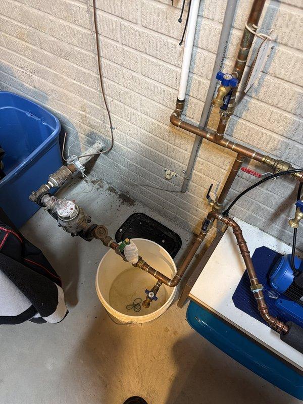 Performed diagnostic evaluation and provided estimate for replacement of aging city pressure booster pump system, approximately 12-13 years old, located in utility area alongside water softener and water heater. Documented existing installation with copper piping connections showing typical age-related patina and corrosion, pump mounted on blue base platform with electrical connections and multiple plumbing ports requiring replacement due to equipment age and wear.