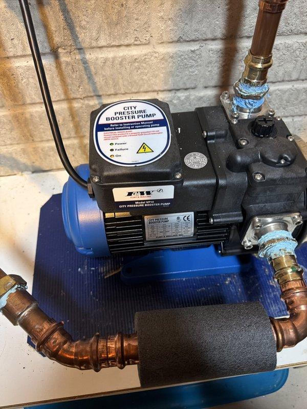 Performed diagnostic evaluation and provided estimate for replacement of aging city pressure booster pump system, approximately 12-13 years old, located in utility area alongside water softener and water heater. Documented existing installation with copper piping connections showing typical age-related patina and corrosion, pump mounted on blue base platform with electrical connections and multiple plumbing ports requiring replacement due to equipment age and wear.