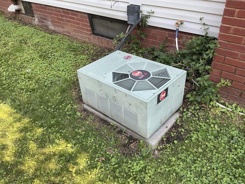 Conducted on-site evaluation for replacement of 31-year-old furnace and air conditioning system. Documented existing Rheem outdoor condenser unit positioned on concrete pad adjacent to brick foundation with white vinyl siding, noting refrigerant line routing and current installation configuration. Photographed equipment placement, surrounding landscape conditions, and utility connections including electrical service and PVC condensate drainage to provide comprehensive documentation for replacement proposal and customer confirmation of work scope.
