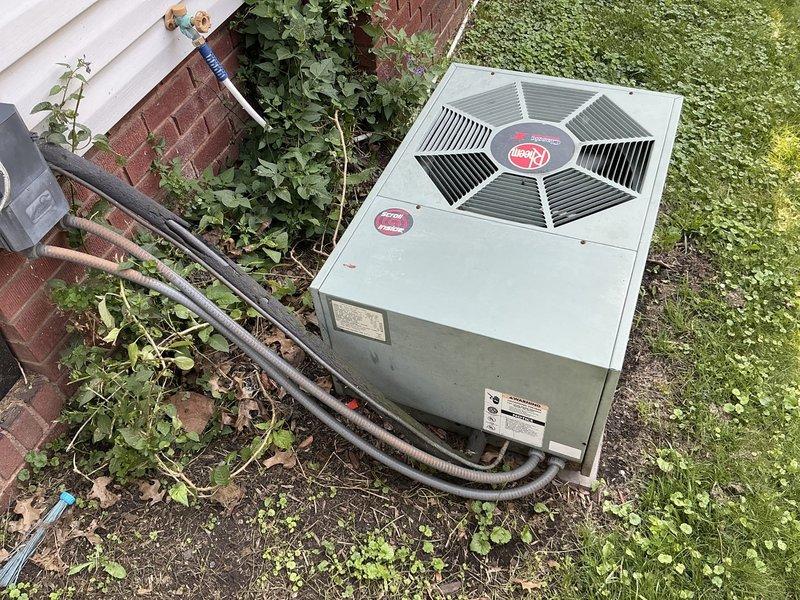 Conducted on-site evaluation for replacement of 31-year-old furnace and air conditioning system. Documented existing Rheem outdoor condenser unit positioned on concrete pad adjacent to brick foundation with white vinyl siding, noting refrigerant line routing and current installation configuration. Photographed equipment placement, surrounding landscape conditions, and utility connections including electrical service and PVC condensate drainage to provide comprehensive documentation for replacement proposal and customer confirmation of work scope.