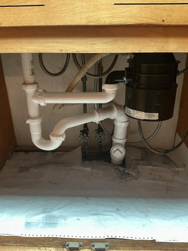 Replaced complete kitchen drain line from under sink cabinet extending down through basement to main stack. Removed existing damaged piping and installed new white PVC drain assembly including P-trap configuration under sink, connecting dual drain lines to garbage disposal unit. Ran new drainage pipe vertically through floor down to basement level, reconnecting to previously broken vent line and tying back into main plumbing stack. All connections properly fitted and secured to ensure code-compliant drainage and venting throughout system.