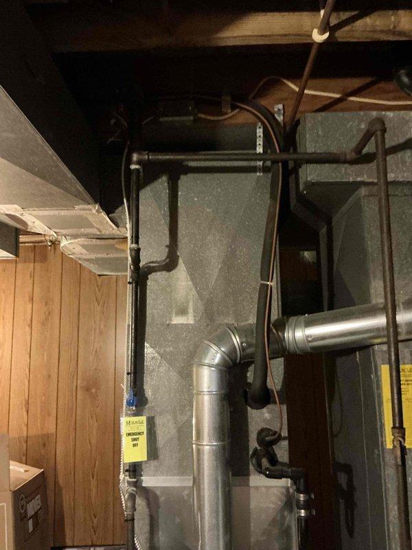 Completed estimate for replacement of 45-year-old air conditioning system. Evaluated existing dual outdoor condensing units positioned on exterior concrete pad against brick building wall, connected via black electrical conduit. Assessed indoor air handler and furnace unit located in basement utility area with existing ductwork and piping infrastructure. Documented current system configuration including galvanized and aluminum ductwork with 90-degree elbows, metal pipe hangers, and ceiling-mounted support structures in unfinished basement space. Provided replacement recommendations for complete HVAC system upgrade.