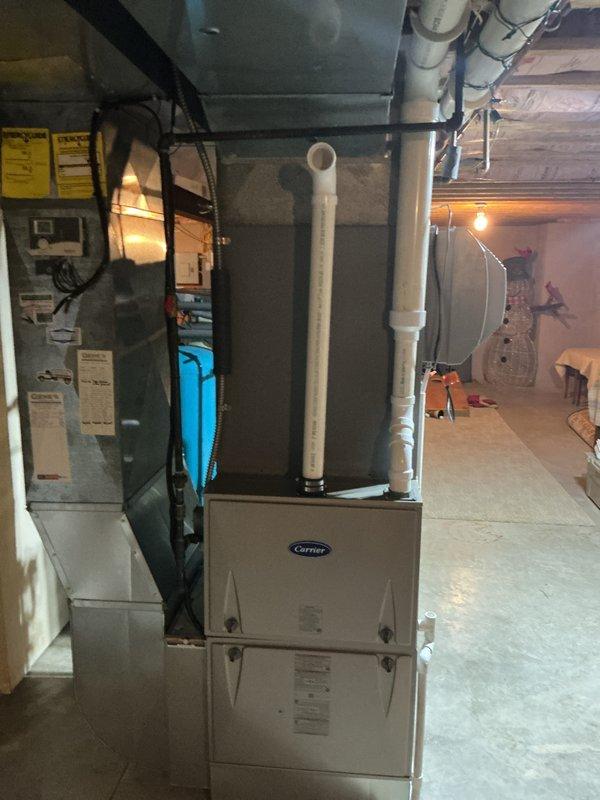 Completed installation of new Carrier HVAC system including furnace, air handler, and outdoor condensing unit. Interior equipment installed in basement with proper venting via white PVC piping, and outdoor AC unit positioned on gravel pad against exterior wall with appropriate electrical disconnect and line set connections. System fully operational with all components properly mounted, leveled, and secured per manufacturer specifications.