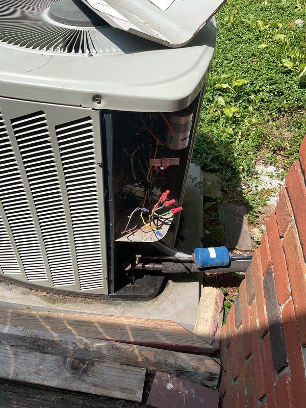 Responded to no cooling complaint on 18-year-old residential air conditioning system. Upon arrival, found outdoor condenser unit with fan motor operating but system failing to produce cooling. Observed vegetation growth obstructing airflow around condenser coils and internal components. Removed access panel to inspect electrical system, including capacitor and wiring connections. Documented condition of unit with photographs showing aged equipment, compromised internal components, and environmental debris accumulation affecting system performance.