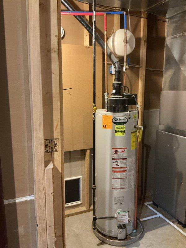 Responded to service call for leaking A.O. Smith tanked water heater with unusual dual-fuel configuration having both gas and electric connections. Conducted thorough assessment of the unit and photographed existing installation showing water heater positioned in unfinished basement with associated plumbing connections, black pump assembly, copper supply lines with color-coded sections, white expansion tank, and pressure relief piping. Documented complete system setup including all pipe routing, fittings, and component placement to provide comprehensive replacement estimate for failed unit.