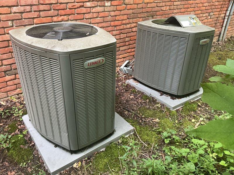 Completed installation of two outdoor Lennox air conditioning condenser units positioned side by side on concrete pads against exterior brick wall, with proper clearance spacing between units and building structure. Indoor coil installation performed in basement mechanical room, integrated with existing furnace system, including proper refrigerant line connections, condensate drain piping with white PVC, and electrical connections to support dual-zone cooling system operation.