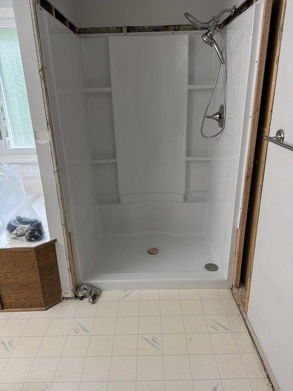 Completed installation of a new 48x36 center drain shower unit with fiberglass surround and integrated shelving recesses. Installed chrome handheld showerhead assembly and dual drain configuration with brass and chrome drain covers. Shower enclosure features molded corner construction with decorative tile border trim at upper perimeter. Unit was set on existing tile floor base with exposed framing prepared for final trim work completion.