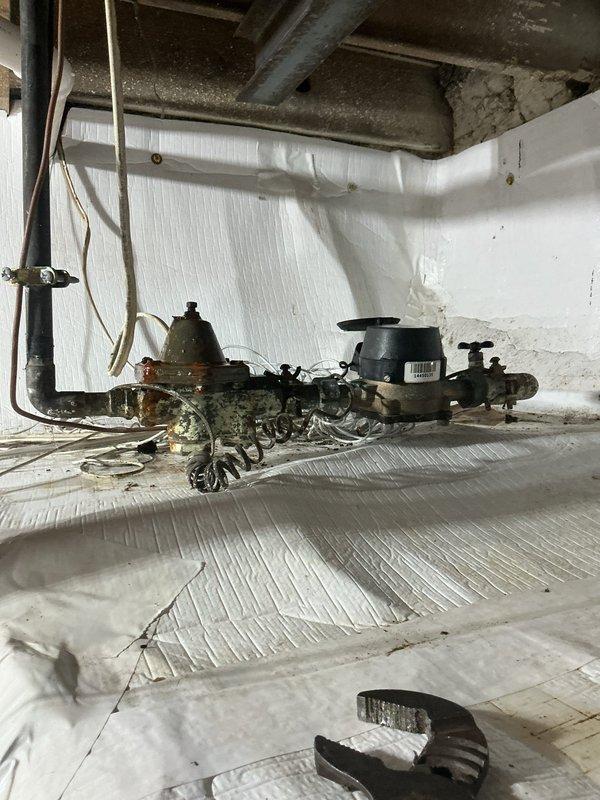 Responded to emergency call for active water line failure in basement. Upon arrival, found water pouring from compromised main line into basement area. Homeowner had already shut off main water supply to stop active flooding. Assessed crawl space and identified severely corroded pump assembly and valve components as source of failure. Inspected surrounding plumbing infrastructure including copper piping, valve assemblies, and pump connections, all showing significant age-related deterioration and corrosion. Documented extensive rust and patina buildup on mechanical components. Evaluated electrical wiring and connections in affected area for water damage. Photographed compromised equipment and surrounding conditions for repair assessment. Recommended main line repair or replacement to restore water service and prevent future failures.