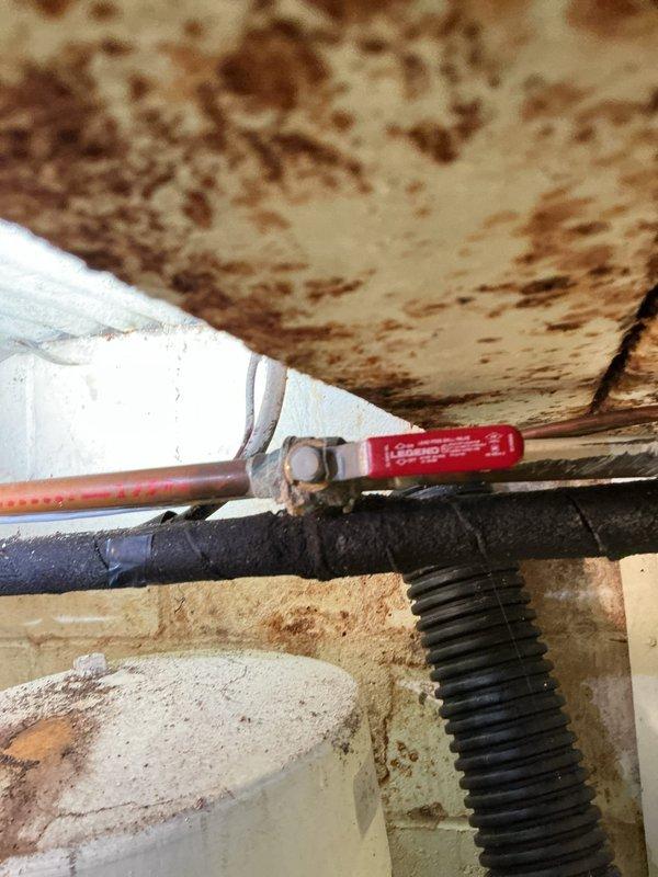 Responded to service call for water leak of unknown origin in residential property. Upon arrival, conducted thorough inspection of basement utility area and identified extensive water damage and rust staining on ceiling surfaces, walls, and mechanical equipment. Located active leak source at water heater connections, with significant moisture intrusion evident from brown discoloration patterns on overhead surfaces and corroded piping components. Observed compromised pipe fittings with visible corrosion on copper supply lines and deteriorated connections at water heater inlet. Documented existing conditions showing heavy rust streaking on white cylindrical water heater tank and surrounding infrastructure, indicating prolonged moisture exposure. Photographed damaged areas including water-stained ceiling joists, corroded piping systems with green-tinted valve fittings, and affected concrete floor surfaces for record purposes.