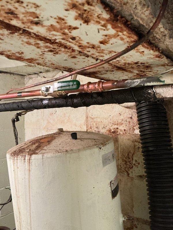 Responded to service call for water leak of unknown origin in residential property. Upon arrival, conducted thorough inspection of basement utility area and identified extensive water damage and rust staining on ceiling surfaces, walls, and mechanical equipment. Located active leak source at water heater connections, with significant moisture intrusion evident from brown discoloration patterns on overhead surfaces and corroded piping components. Observed compromised pipe fittings with visible corrosion on copper supply lines and deteriorated connections at water heater inlet. Documented existing conditions showing heavy rust streaking on white cylindrical water heater tank and surrounding infrastructure, indicating prolonged moisture exposure. Photographed damaged areas including water-stained ceiling joists, corroded piping systems with green-tinted valve fittings, and affected concrete floor surfaces for record purposes.