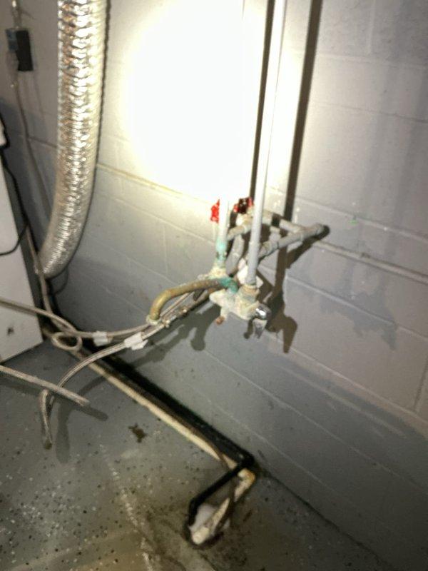 # Basement Floor Drain Backup - Clogged Drain Clearing

Responded to a basement floor drain backup in a 67-year-old residential property. Upon arrival, inspected the utility area and identified the affected floor drain system with visible water staining on the concrete floor indicating recent backup issues.

Accessed the basement plumbing infrastructure, which consisted of older copper supply lines with red and green valve handles, along with horizontal drain piping showing age-related discoloration and mineral deposits. The complex network of pipe connections and fittings displayed typical wear patterns consistent with the home's age.

Located and opened the floor drain access point, revealing a cylindrical underground drainage structure with deteriorated green coating on interior walls. Inspection showed significant accumulation of dark water, debris, and sediment at the bottom of the tank, along with waste material deposits on the liquid surface. The interior walls exhibited water s