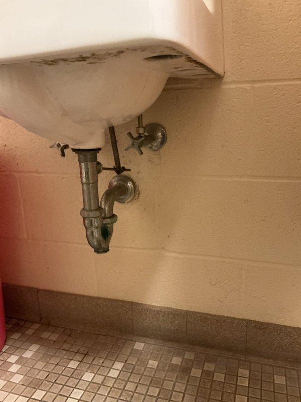 Responded to service call for bathroom fixture assessment in commercial restroom facility. Evaluated existing wall-mounted sink with three-handle faucet configuration, inspecting visible supply lines and chrome P-trap assembly for potential replacement. Assessed urinal flush valve system requiring new flush kit installation. Documented garbage disposal requirement for estimate preparation. Noted visible water damage and staining at tile base along wall perimeter, indicating potential moisture intrusion issues. Photographed existing fixture conditions, plumbing connections, and deteriorated areas to support comprehensive estimate for fixture replacements and necessary repairs.