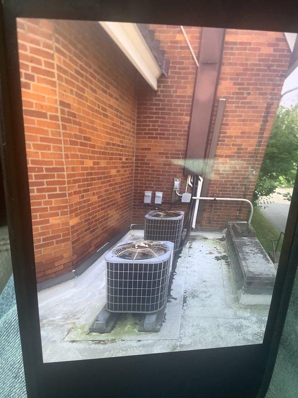 Conducted comprehensive assessment of commercial cooling system including rooftop air conditioning units and air handlers located in attic space above sanctuary area. Both rooftop AC units evaluated and determined to require full replacement due to operational failure and end-of-service-life conditions. Exterior condenser units inspected, showing significant age-related deterioration with visible weathering on concrete pad and surrounding infrastructure. Documented existing refrigerant line sets, electrical disconnects, and PVC piping configuration mounted on exterior brick wall for replacement planning. Photographed current equipment installation including dual condenser setup on concrete pad with associated electrical components and refrigerant piping. Prepared detailed estimate for complete system replacement of both rooftop units and coordinated ladder access requirements for attic air handler inspection and future installation work.