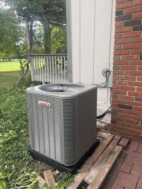 Completed installation of Lennox air conditioning system with matching evaporator coil at residential property. Unit positioned on wooden platform base adjacent to brick and vinyl siding exterior, with proper electrical connections including 30-amp fuses and conduit routing to outdoor disconnect. All refrigerant lines connected, system tested, and confirmed operational.