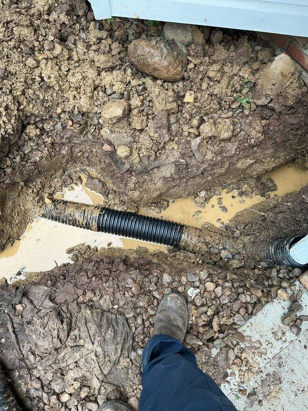 Responded to emergency call for damaged downspout caused by electrical contractor impact. Upon arrival, excavated area around building foundation to expose the damaged black corrugated drainage pipe that had been struck during electrical work. Removed damaged section of downspout drain line, installed replacement corrugated drainage pipe, and properly backfilled excavation with gravel and soil to restore proper drainage function and prevent water accumulation near foundation.