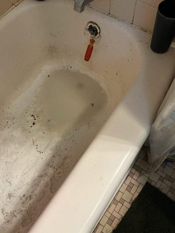 Responded to upstairs bathtub drainage issue. Upon arrival, found bathtub with standing water and damaged drain assembly with red rubber component protruding from drain opening. Accessed plumbing system through floor access panel to inspect drain line and shutoff valves. Removed defective drain stopper mechanism and cleared obstruction from drain line. Tested drainage flow and verified proper operation of bathtub drain system.