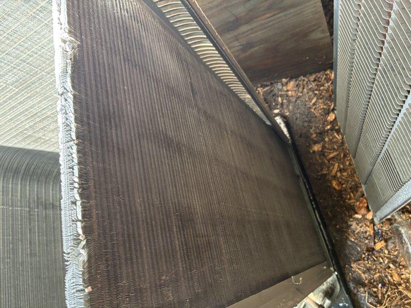 Completed MVP cooling maintenance visit on residential HVAC system. Upon inspection, discovered significant debris accumulation between evaporator coil assemblies, including dead leaves, dirt, and organic matter concentrated in the lower section where coil units meet. Observed heavy dirt and dust buildup covering substantial portions of the outdoor condenser unit coils, with visible discoloration across center and right sections contrasting with cleaner metallic fins on left side. Documented deterioration along coil edge with fraying material present. Unit showed signs of weathering and required cleaning to restore optimal heat exchange efficiency and system performance.